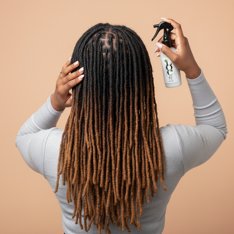 Person with long braided hair holding a spray bottle against a beige background